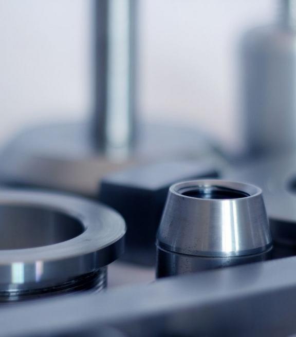 Nahaufnahme mehrerer gedrehter Metallteile aus Edelstahl, darunter Ringe, Zylinder und eine Hülse mit Innengewinde, auf einer metallischen Ablage.