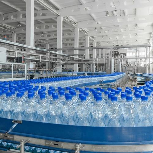 Förderband in einer hellen Produktionshalle, auf dem zahlreiche mit Wasser gefüllte Plastikflaschen mit blauen Verschlüssen dicht an dicht transportiert werden.