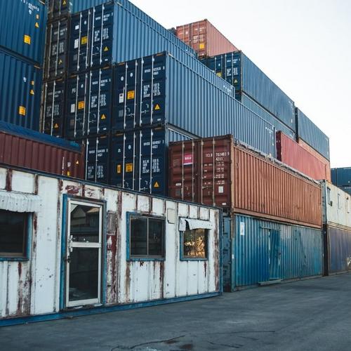 Gestapelte Schiffscontainer in Blau, Rot und Grau auf einem Containerterminal, davor ein flaches Gebäude mit Fenstern.